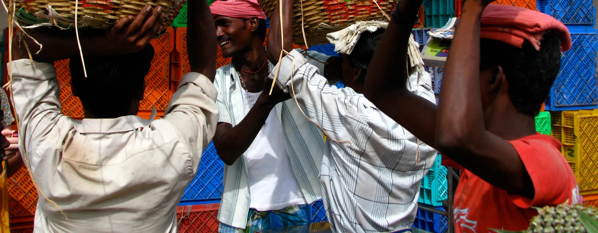 Market labourers carry large woven baskets among stacked produce crates at Chennai’s Koyambedu wholesale market.