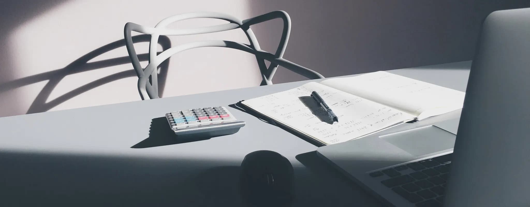 A calculator, handwritten figures and an open laptop arranged on a sunlit desk.