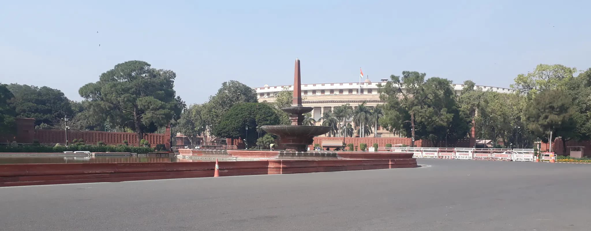 The circular Parliament House in New Delhi behind a red sandstone fountain and an empty roadway.