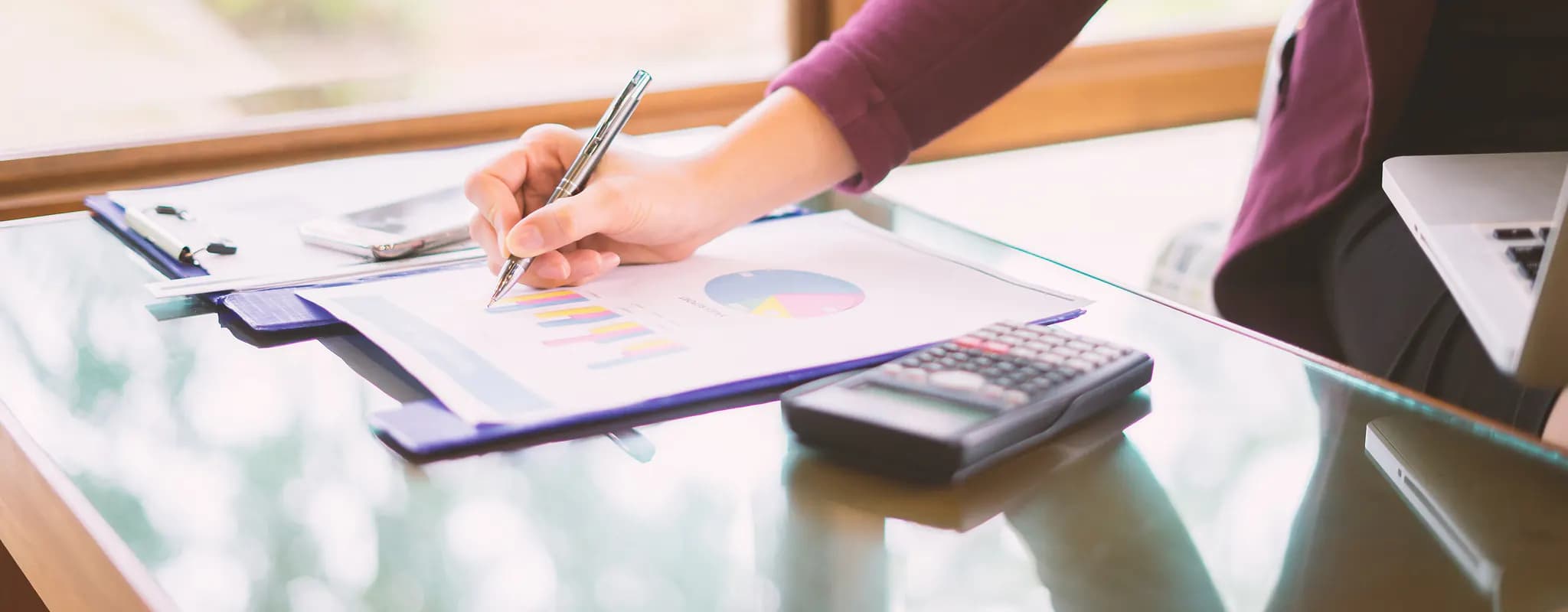 A businesswoman reviews financial charts at a glass desk beside a calculator and laptop.