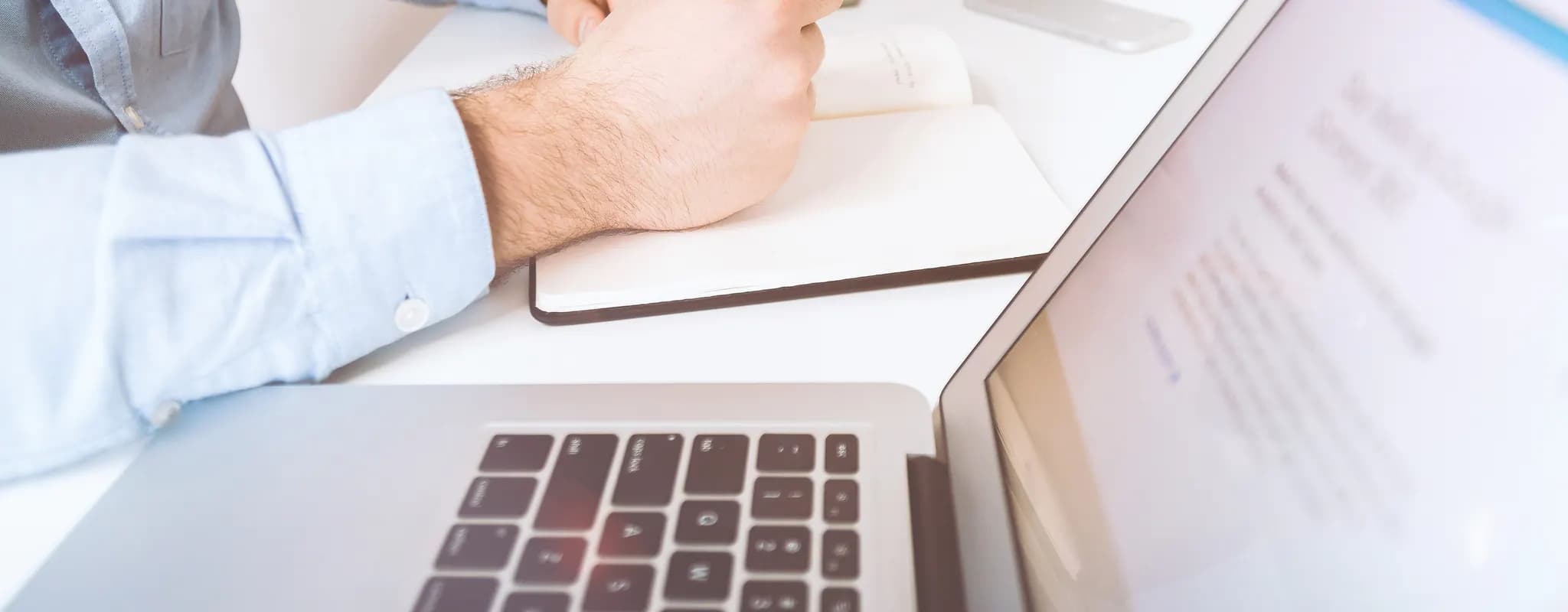 A professional writes notes beside an open laptop at a bright office desk.