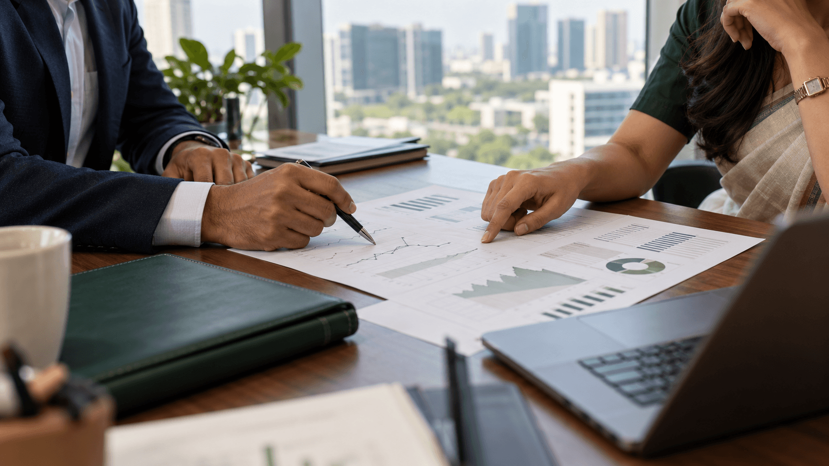 Two professionals reviewing financial charts and reports beside a laptop in a sunlit office.