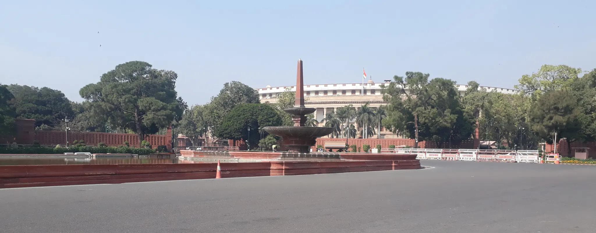 The circular Parliament House in New Delhi behind a red sandstone fountain and an empty roadway.