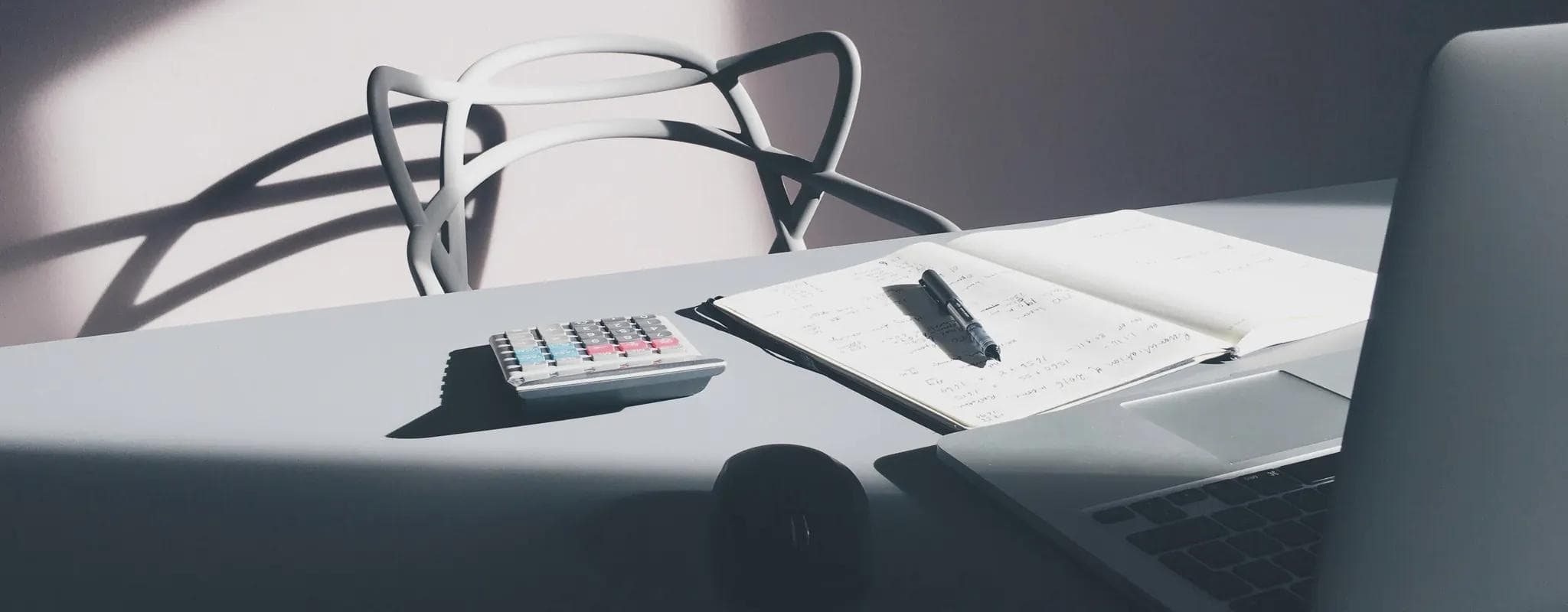 A calculator, handwritten figures and an open laptop arranged on a sunlit desk.