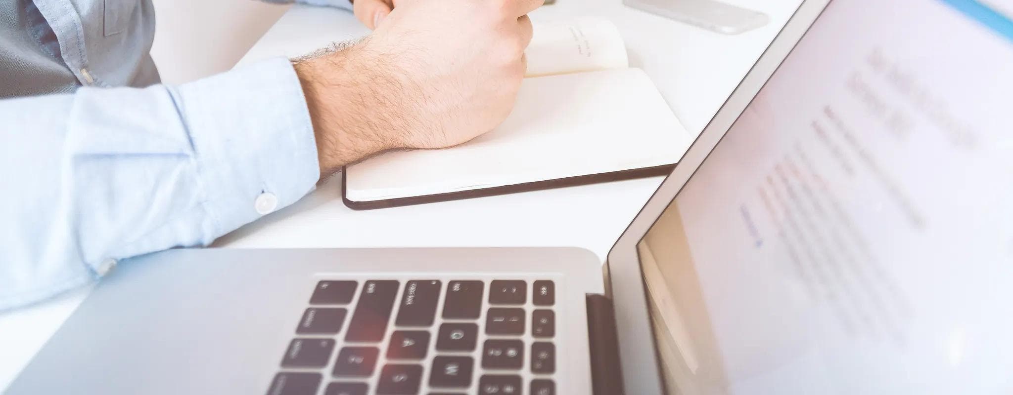 A professional writes notes beside an open laptop at a bright office desk.